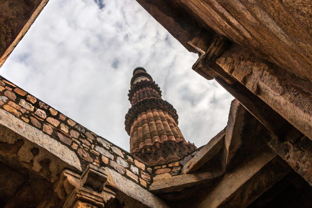 A captivating low-angle view of Qutub Minar tower in Delhi, India, surrounded by rustic stone walls under a cloudy sky.